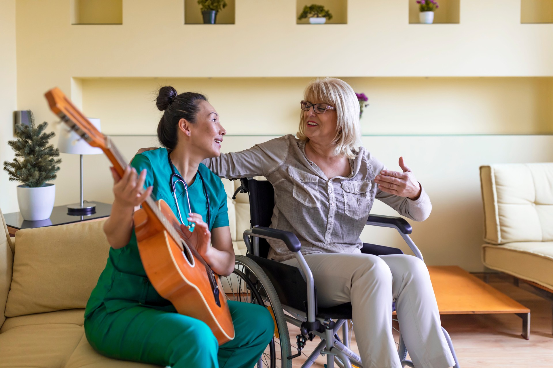 Female Home Caregiver is Playing Guitar to an Older Woman in Wheelchair.